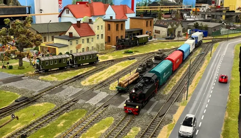 Miniaturowa kolejka z parowozem i wagonami, jak w Energylandii, przemierza malowniczy krajobraz z domkami i drogą.