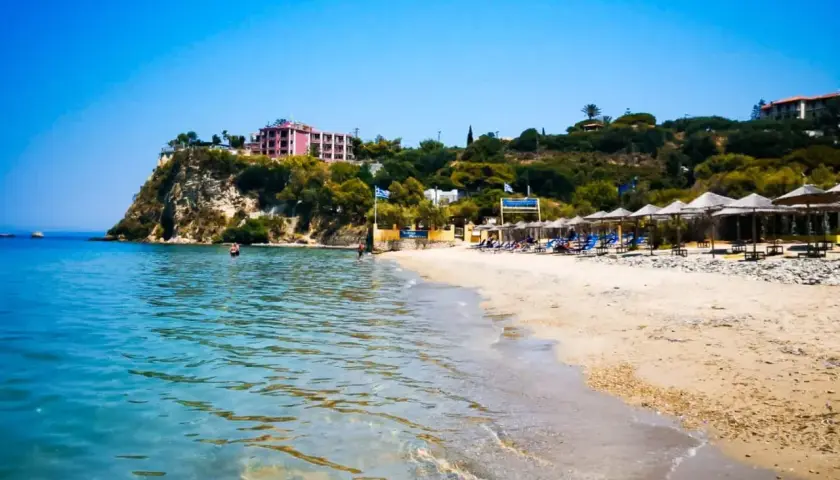 Słoneczny dzień na plaży Alykanas. Turkusowa woda, piaszczysty brzeg z leżakami i parasolami, a w oddali hotel na klifie.