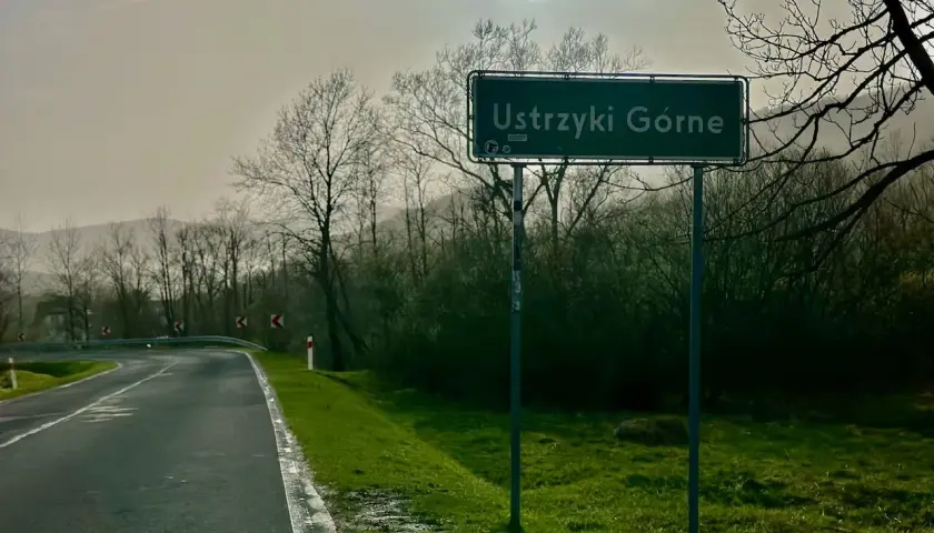 Droga z Krakowa do Ustrzyk Górnych. Znak z nazwą miejscowości Ustrzyki Górne na tle drzew i mglistego krajobrazu.