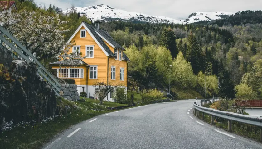Wjazd do Norwegii. Żółty dom z białymi oknami, kwitnące drzewo, droga wijąca się wśród zieleni i ośnieżone góry w tle.