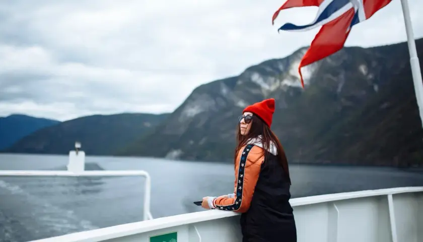 Kobieta na statku pod norweską flagą podziwia fiordy. Podróż po Norwegii, mimo że piękna, może wiązać się z wysokimi cenami.