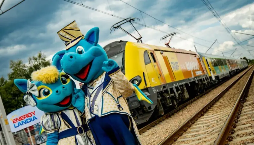 Dwa niebieskie smoki w strojach, obok znaku Energylandia, pozdrawiają przed pociągiem RMF. Idealny dojazd do Energylandii!