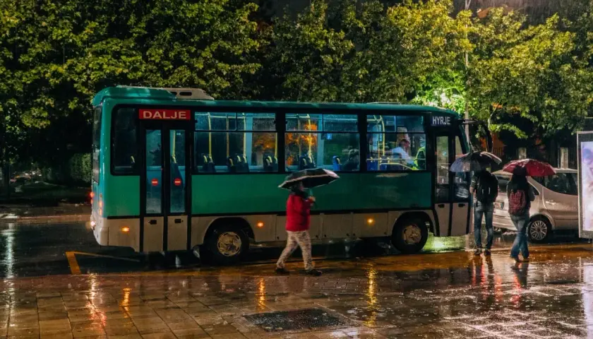 Deszczowa noc w Tirana Durres. Autobus z napisem "Dalje" i "Hyrje" stoi na mokrej ulicy, ludzie z parasolami czekają.