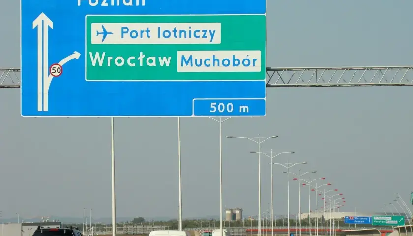 Znak drogowy pokazuje, jak dojechać na lotnisko we Wrocławiu z dworca PKP. Kierunek na lotnisko i Wrocław Muchobór jest po prawej stronie.