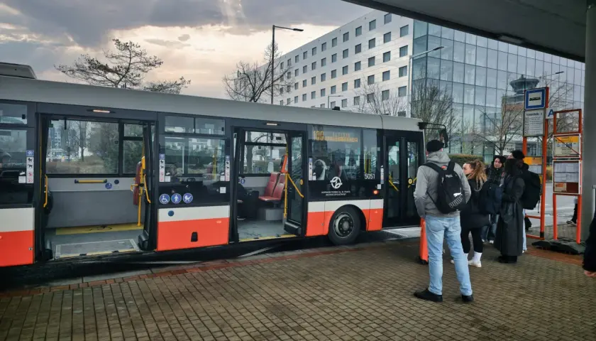 Praga autobus czeka na pasażerów przy nowoczesnym budynku. Grupa ludzi stoi na przystanku, gotowa do podróży.
