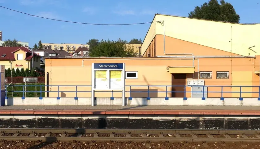Peron kolejowy w Starachowicach z tablicą informacyjną i budynkiem dworca w tle.