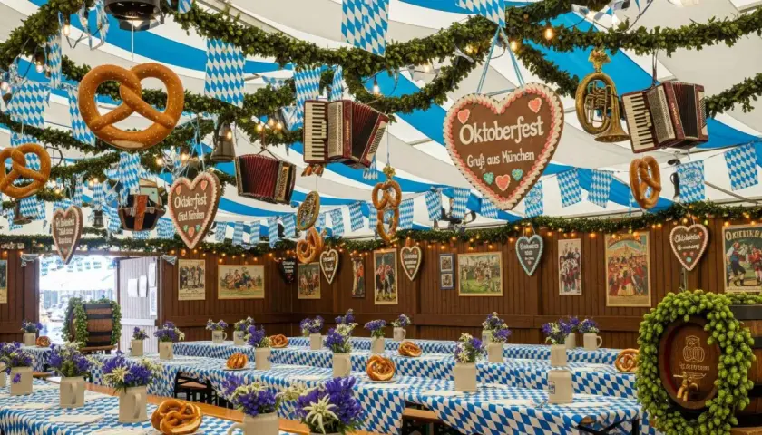 Wystrój namiotu na Oktoberfest w Monachium: wiszące precle, akordeony, serca z piernika i niebiesko-białe flagi. Stoły przykryte kratkowanymi obrusami, ozdobione kwiatami i beczką chmielu.