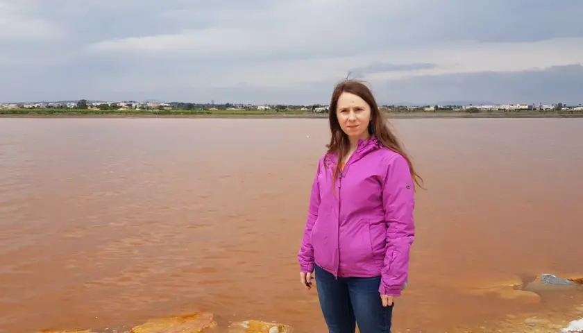 Kobieta w różowej kurtce stoi nad brzegiem Alicante różowego jeziora. W tle widać miasto.
