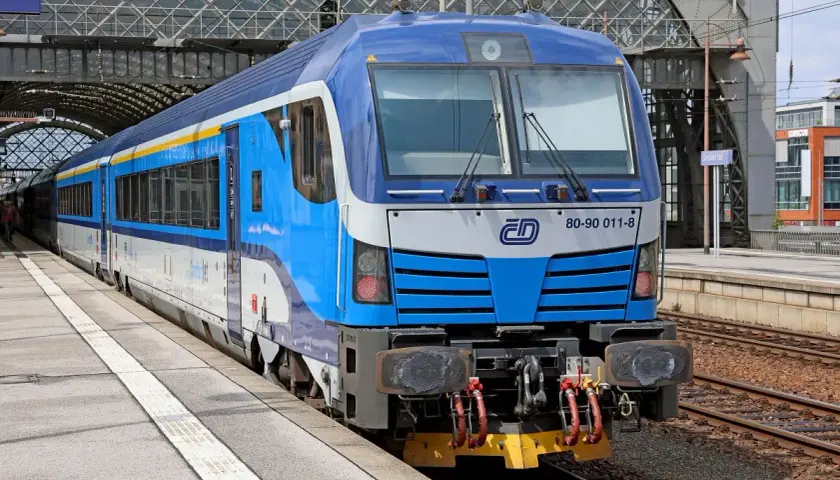 Niebiesko-biały pociąg z żółtym pasem, gotowy do podróży do Czech, stoi na peronie pod nowoczesnym zadaszeniem dworca.
