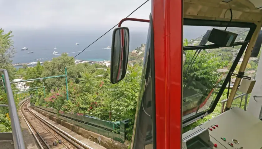Czerwony wagonik kolejki linowej zjeżdża po stromym torze, oferując widok na zatokę pełną jachtów. To podróż jak z Neapolu do Sorrento, pełna zieleni i błękitu.