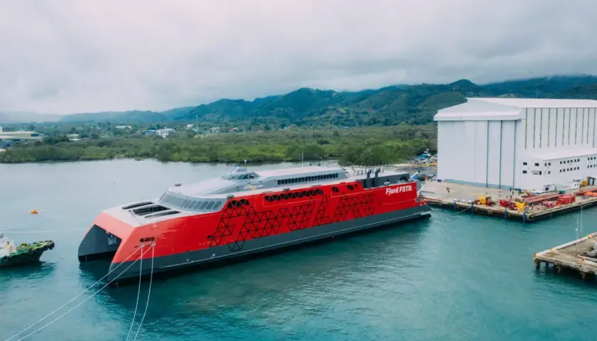 Nowy, czerwono-szary prom Fjord FSTR gotowy do rejsu do Norwegii. W tle zielone wzgórza i biała hala produkcyjna.