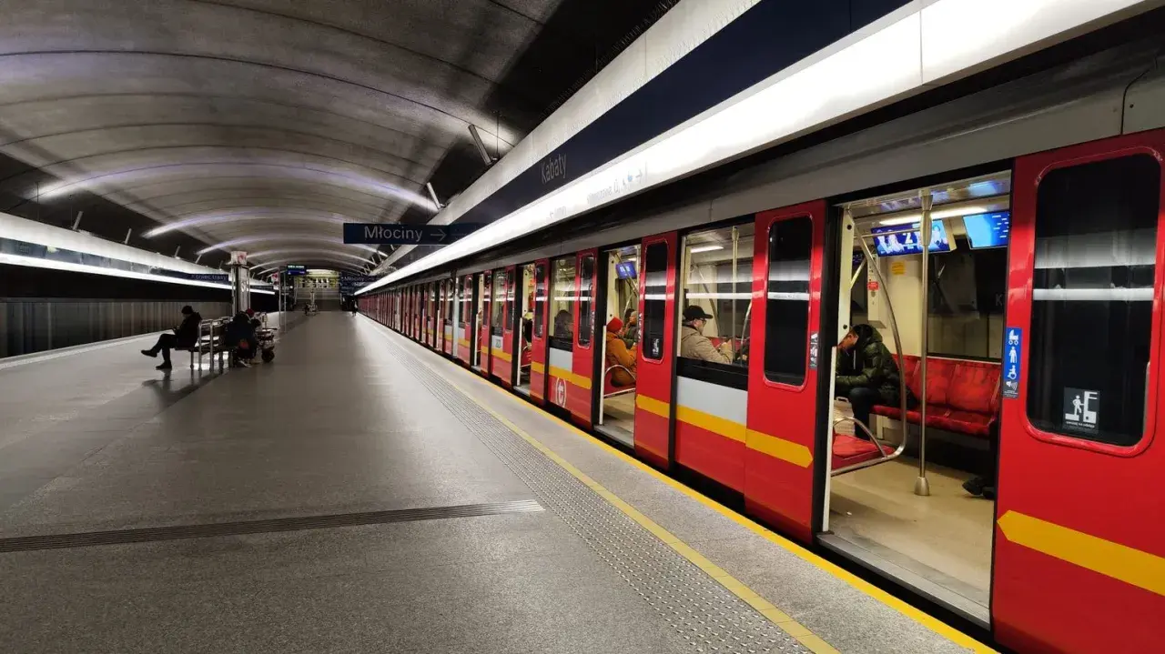 Stacja metra Młociny, do której jeździ metro w Warszawie. Czerwony pociąg z żółtym pasem stoi na peronie, czekając na pasażerów.