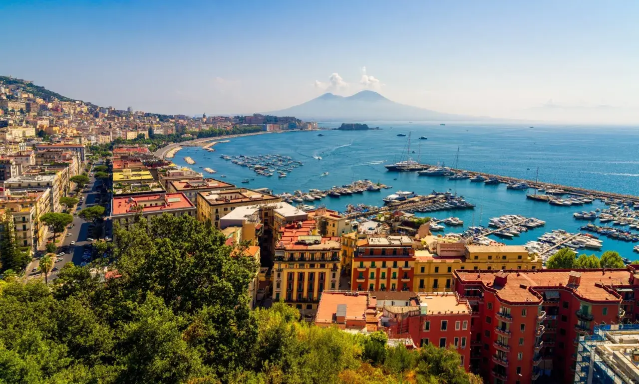 Widok na Zatokę Neapolitańską z Wezuwiuszem w tle. Miasto rozciąga się wzdłuż wybrzeża, a port pełen jest łodzi. Roma Napoli to marzenie wielu podróżników.