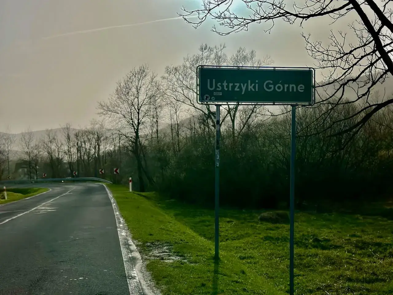 Znak drogowy "Ustrzyki Górne" na tle mglistego krajobrazu z krętą drogą i drzewami.
