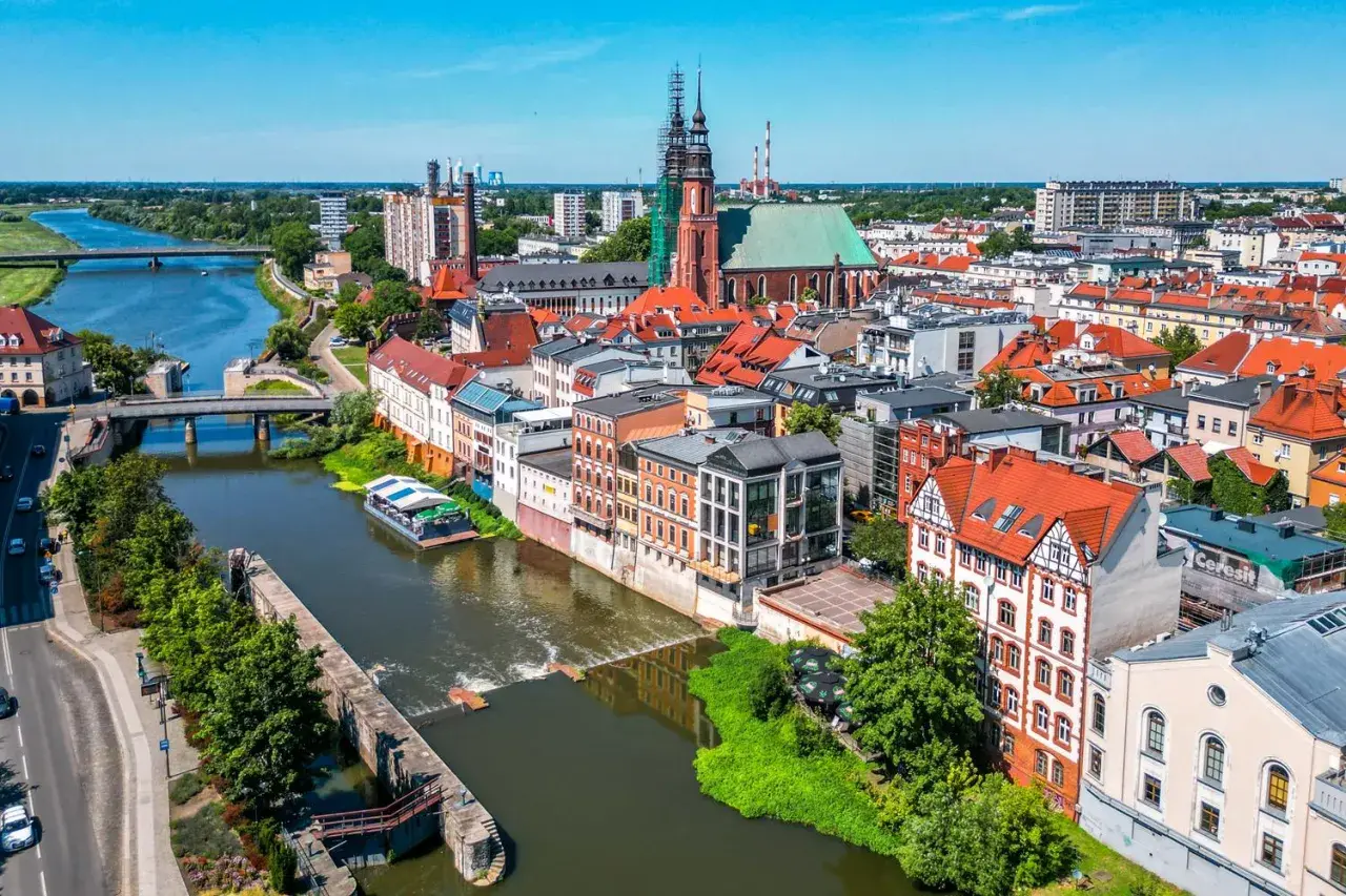 Widok na Opole z rzeką i grodem. Słoneczny dzień nad Odrą, widać kościół i kamienice.