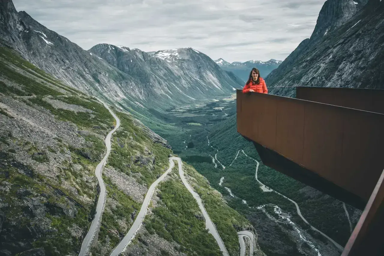 Kobieta na platformie widokowej podziwia serpentyny drogi Trollstigen w otoczeniu gór.