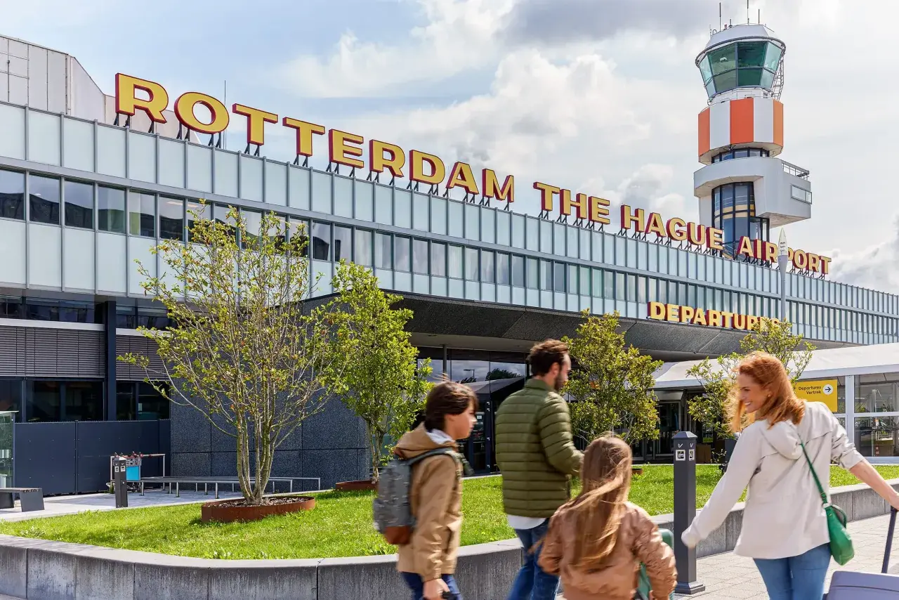 Rodzina zmierza na lotnisko Rotterdam The Hague Airport. W tle widoczna wieża kontroli lotów i napis z nazwą lotniska.