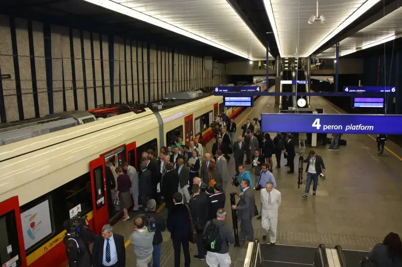 Ludzie wsiadają do pociągu na stacji Warszawa Centralna. Tablica informuje o peronie 4, który obsługuje połączenie do Lotniska Chopina.