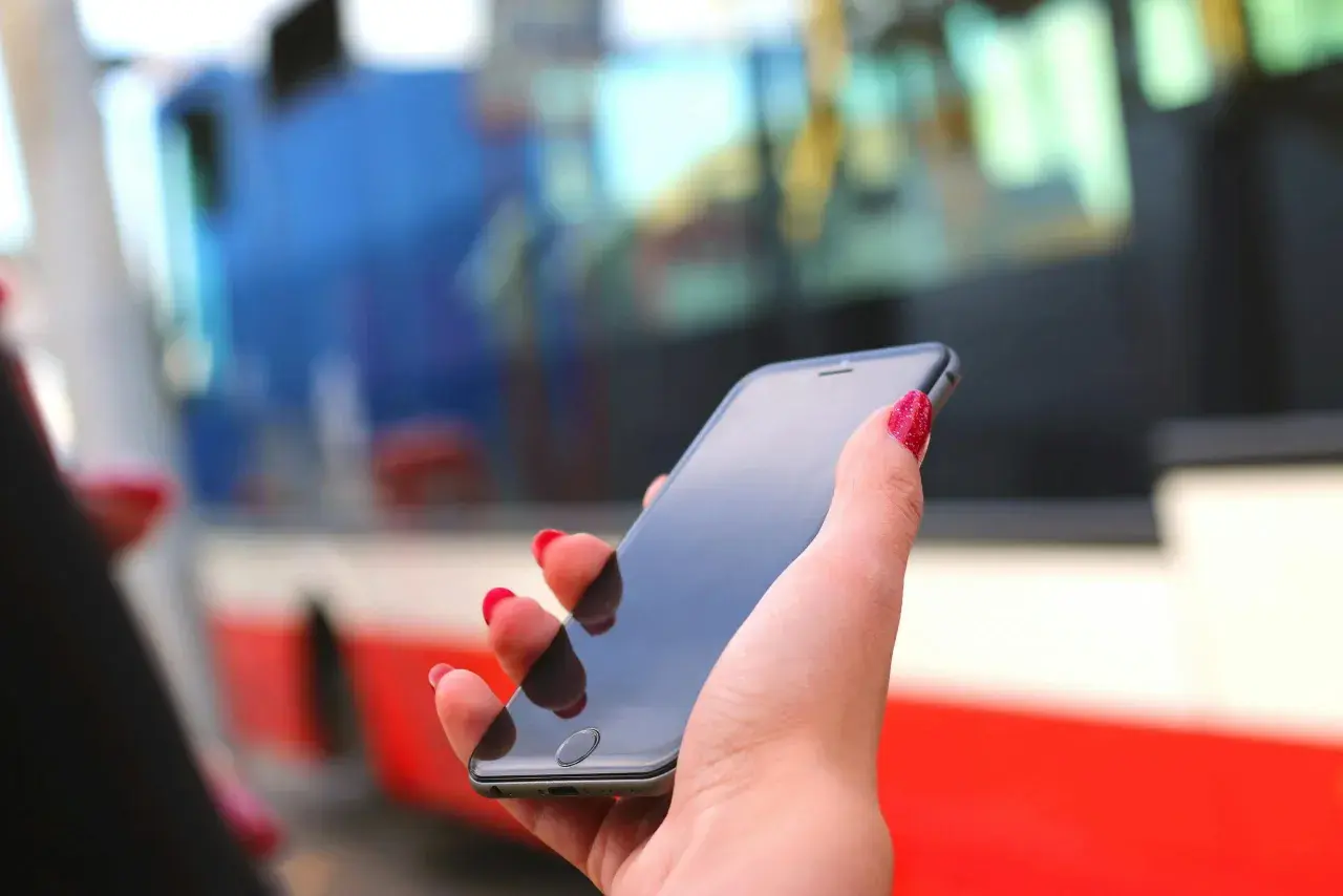 Dłoń z czerwonymi paznokciami trzyma smartfon, na którym można kupić bilet ZTM Warszawa online. W tle widać autobus.