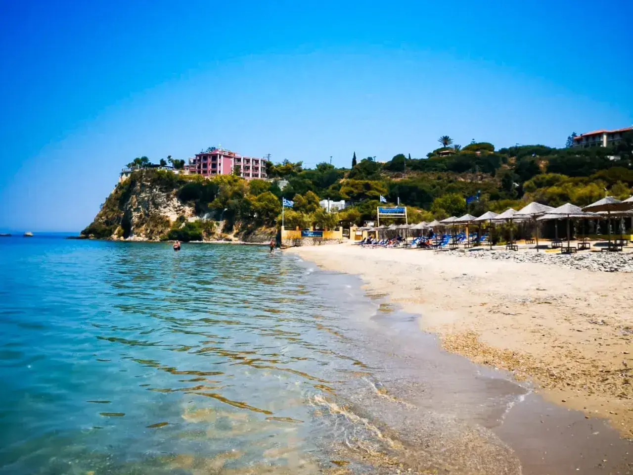 Słoneczny dzień na plaży Alykanas. Turkusowa woda, piaszczysty brzeg z leżakami i parasolami, a w oddali hotel na klifie.