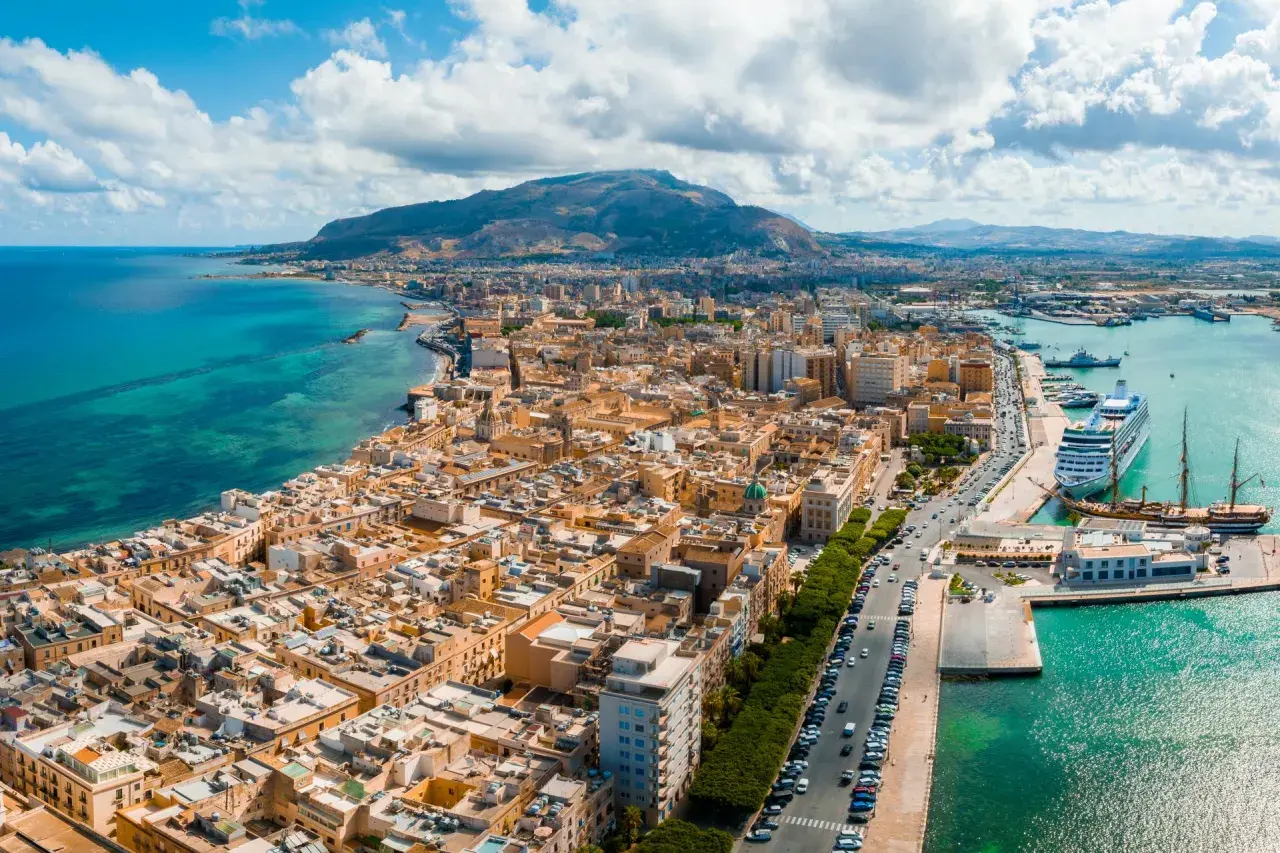Widok na Trapani z lotniska: miasto, port z wycieczkowcem i żaglowcem, góry w tle.