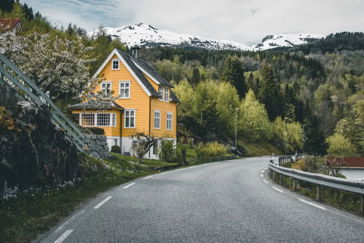 Wjazd do Norwegii. Żółty dom z białymi oknami, kwitnące drzewo, droga wijąca się wśród zieleni i ośnieżone góry w tle.