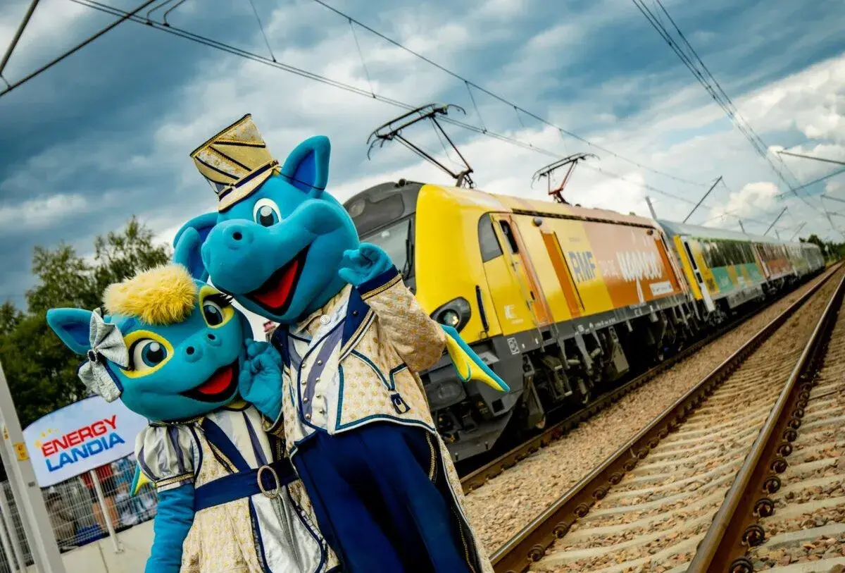 Dwa niebieskie smoki w strojach, obok znaku Energylandia, pozdrawiają przed pociągiem RMF. Idealny dojazd do Energylandii!