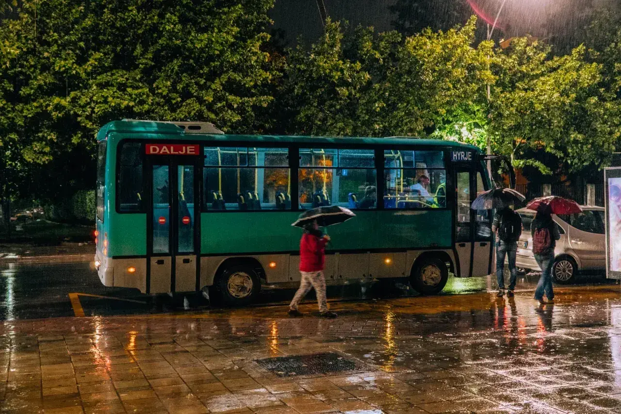 Deszczowa noc w Tirana Durres. Autobus z napisem "Dalje" i "Hyrje" stoi na mokrej ulicy, ludzie z parasolami czekają.