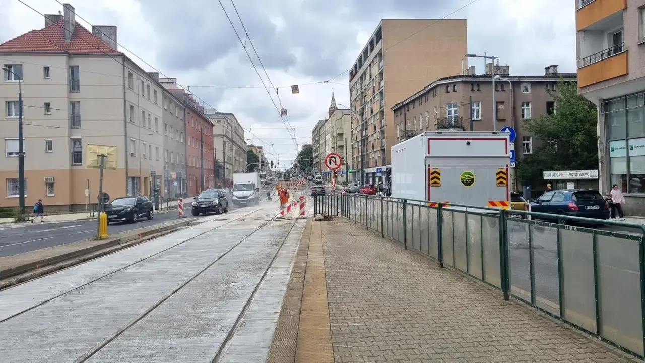 Ulica z torami tramwajowymi w remoncie, budynki mieszkalne, samochody i pracownicy w pomarańczowych kamizelkach.