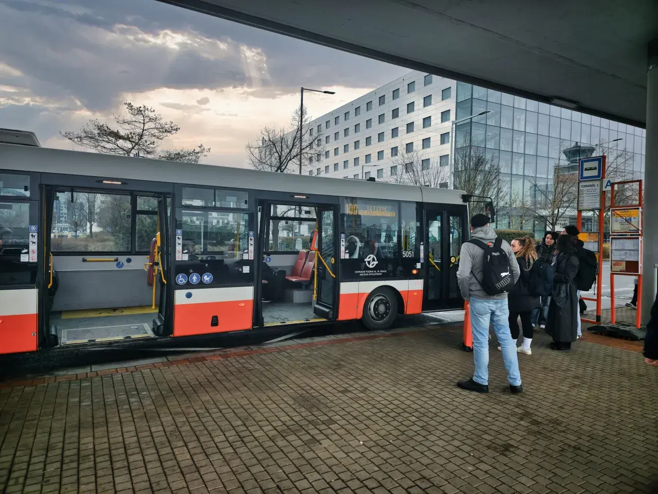 Praga autobus czeka na pasażerów przy nowoczesnym budynku. Grupa ludzi stoi na przystanku, gotowa do podróży.