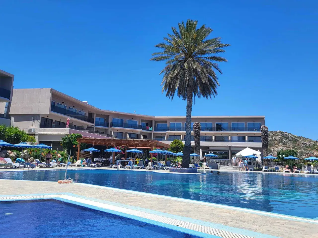 Basen i taras hotelu Cathrin Hotel Rodos z palmami i leżakami pod niebieskimi parasolami.