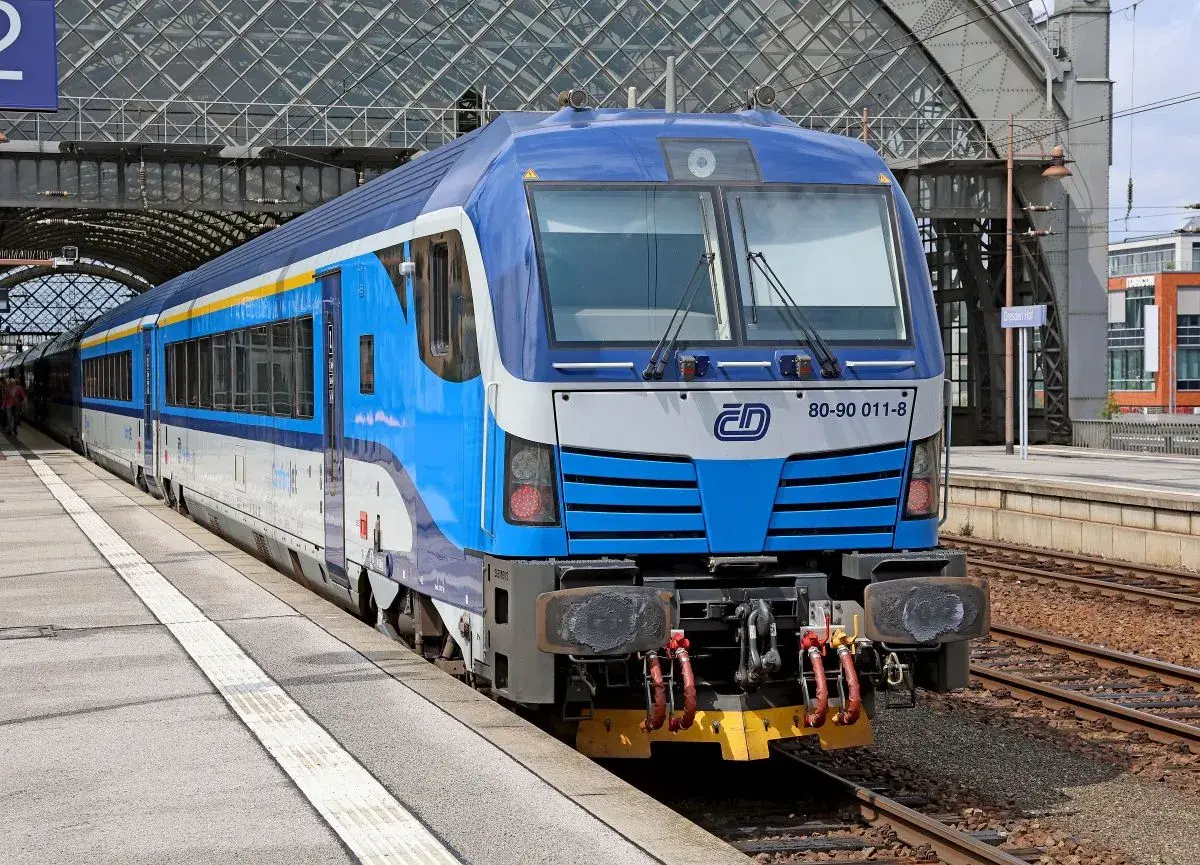 Niebiesko-biały pociąg z żółtym pasem, gotowy do podróży do Czech, stoi na peronie pod nowoczesnym zadaszeniem dworca.