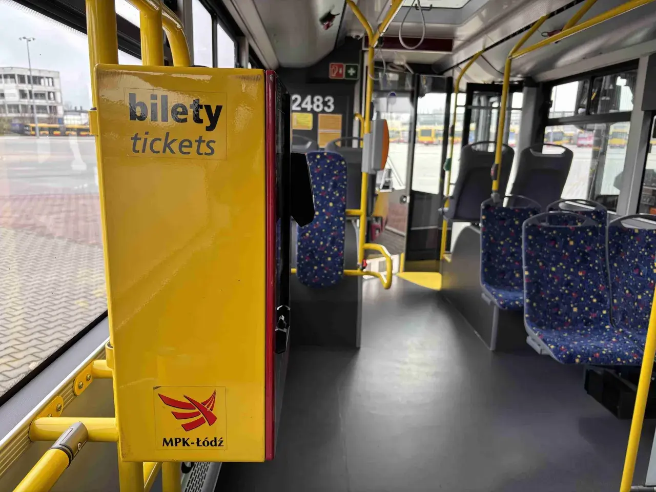 Wnętrze autobusu MPK-Łódź. Żółta skrzynka z napisem "bilety tickets" i logo MPK-Łódź. Widoczne siedzenia i poręcze.