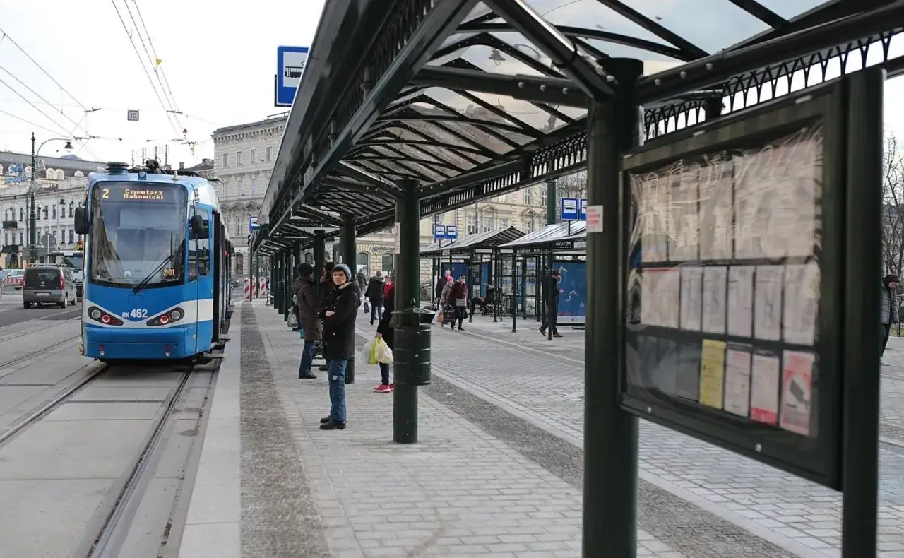 Kraków Swoszowice dojazd: Niebieski tramwaj linii 2 czeka na pasażerów na przystanku. Ludzie stoją pod wiatami, sprawdzając rozkłady jazdy.