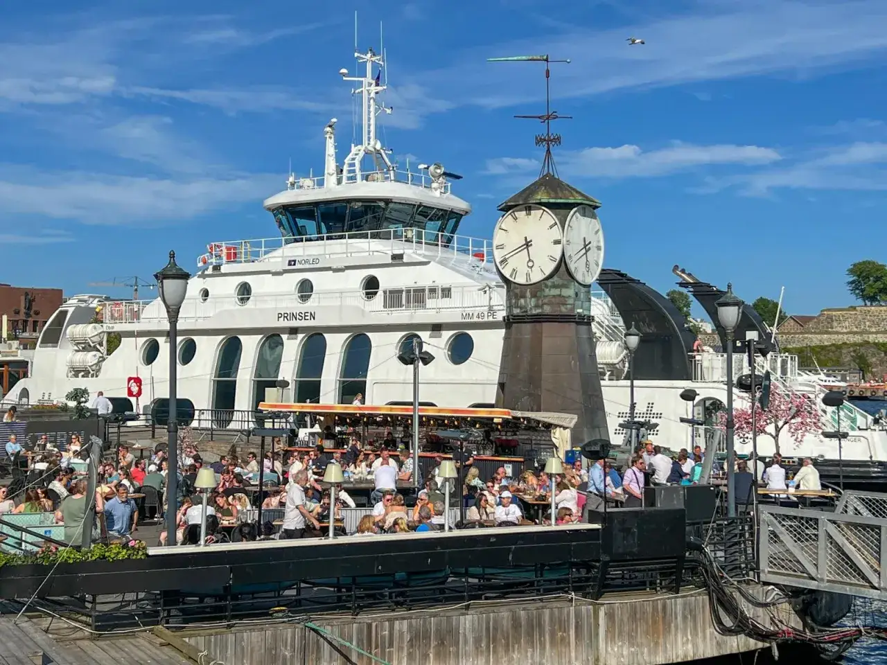 Tłum ludzi w kawiarni nad wodą w Oslo, z widokiem na statek i zegar. Ceny w Oslo mogą być wysokie, ale widok jest bezcenny.