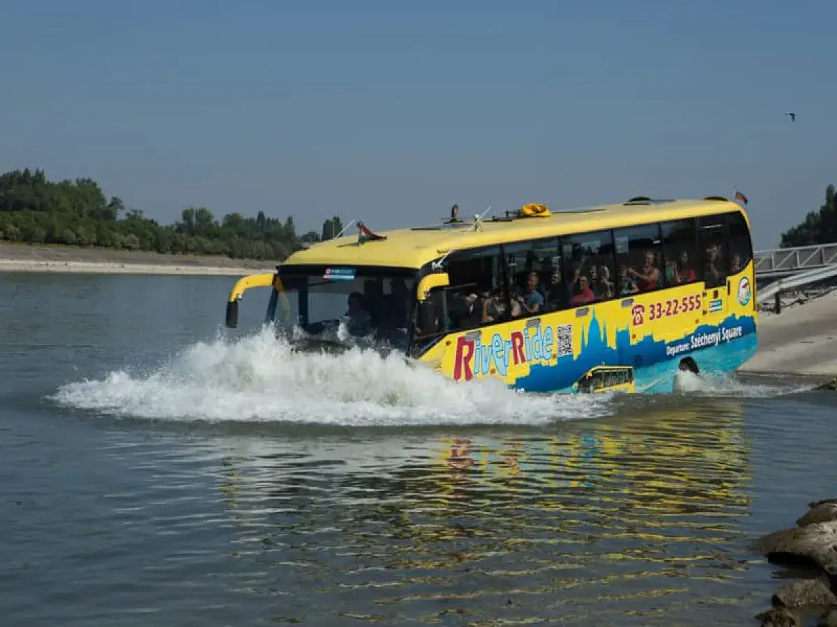 Żółty autobus River Ride wjeżdża do wody w Budapeszcie, tworząc fale. Turyści oglądają widoki.