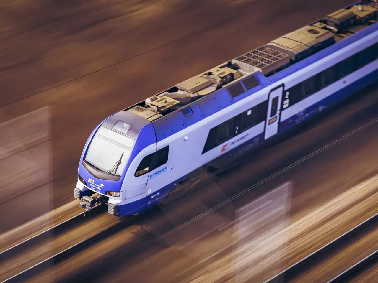 Niebiesko-biały pociąg Stadler mknie po torach, tworząc wrażenie przebookowania krajobrazu.