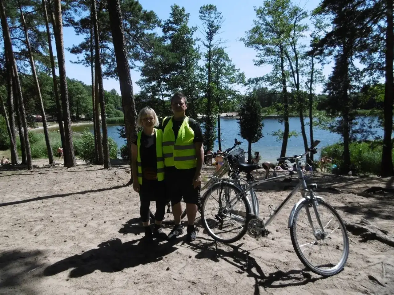 Para rowerzystów w kamizelkach odblaskowych pozuje przy rowerach nad jeziorem, w tle plaża i las. Idealne miejsce na wycieczkę rowerową w okolicach Piecki Bydgoszcz.