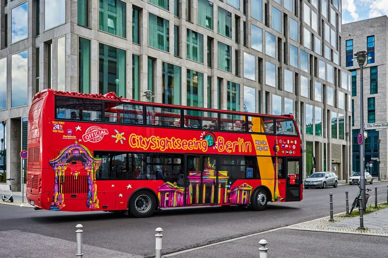 Czerwony, piętrowy autobus City Sightseeing Berlin z kolorowymi grafikami zabytków przejeżdża obok nowoczesnego budynku.