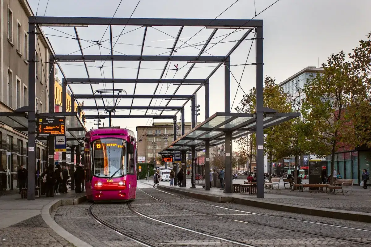 Różowy tramwaj na przystanku w Katowicach. Czekający pasażerowie, tablice informacyjne i nowoczesna architektura. Idealne miejsce, by kupić bilety Katowice i rozpocząć podróż.