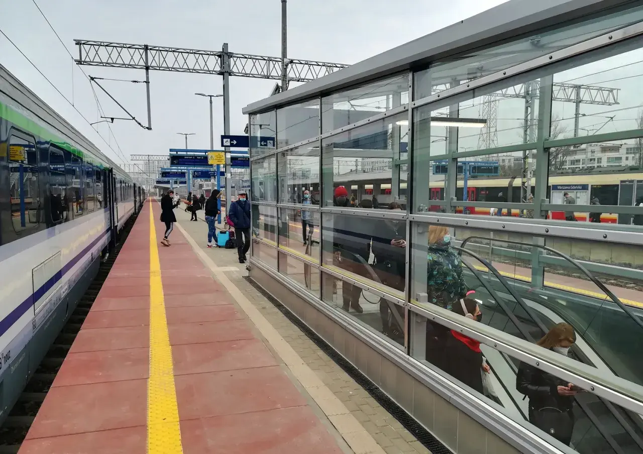 Pociąg Warszawa - Gdańsk czeka na pasażerów. Na peronie widać ludzi z bagażami, a w tle nowoczesna hala z ruchomymi schodami.