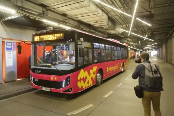 Różowy autobus linii AP3 z napisem "Pyrzowice Port Lotniczy" wjeżdża na pętlę. Obok stoi fotograf z aparatem. Bilety Katowice można kupić na pokładzie.