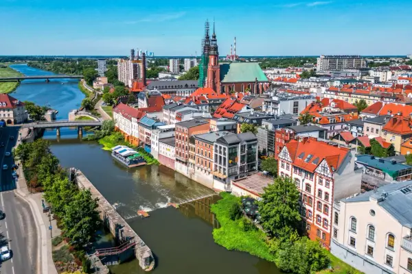 Widok na Opole z rzeką i grodem. Słoneczny dzień nad Odrą, widać kościół i kamienice.