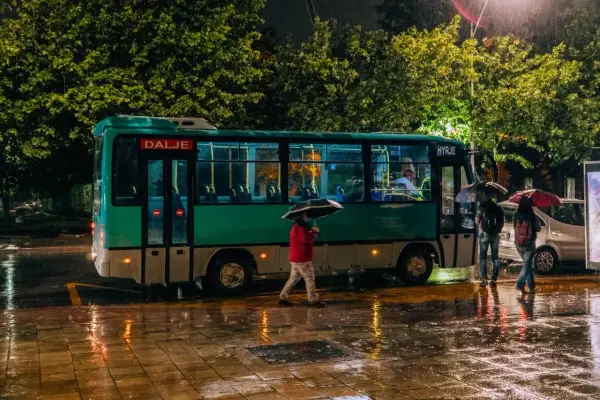 Deszczowa noc w Tirana Durres. Autobus z napisem "Dalje" i "Hyrje" stoi na mokrej ulicy, ludzie z parasolami czekają.